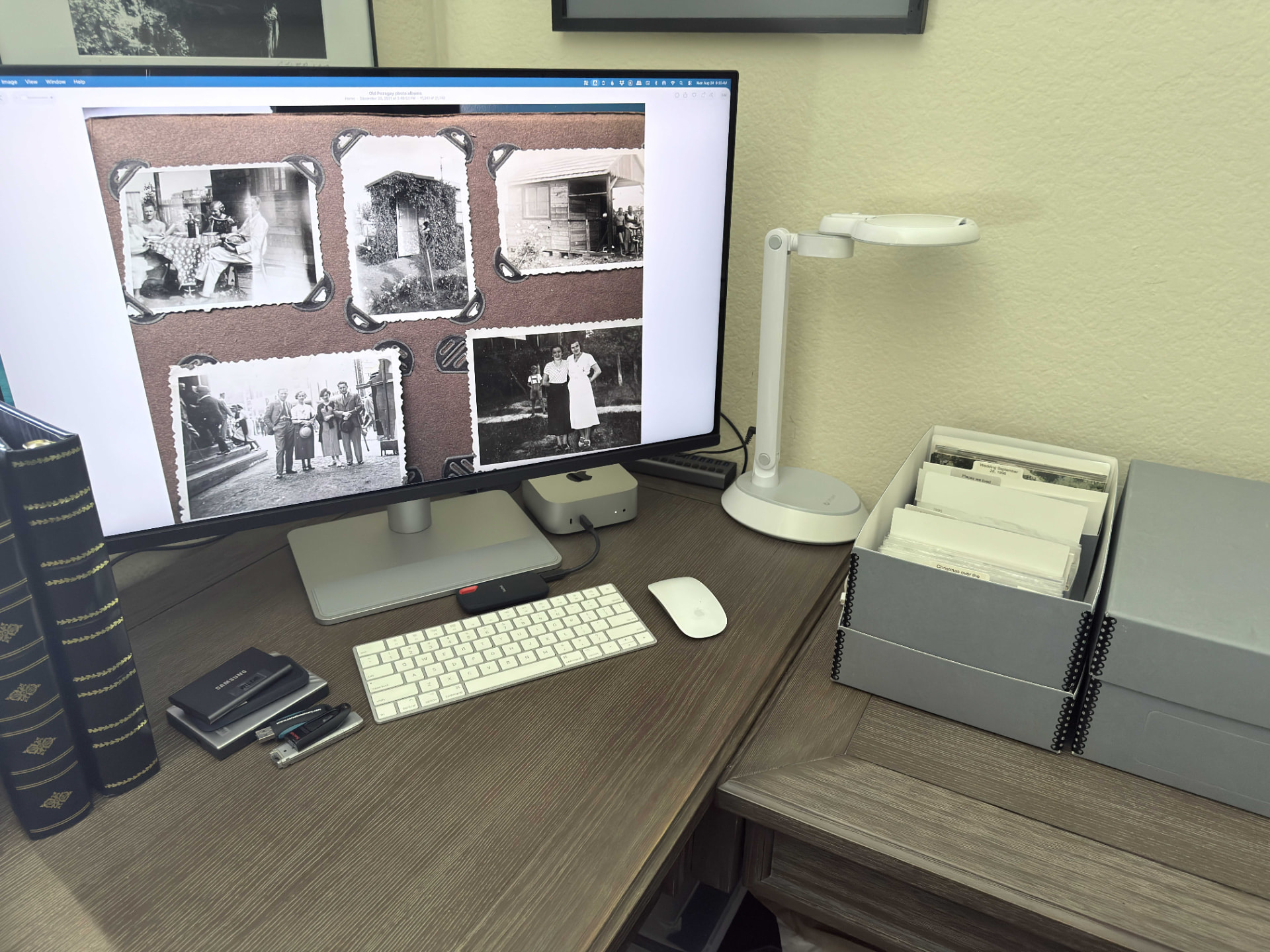 A photo organizer's desk showing digitized vintage family photos on a monitor alongside archival storage boxes